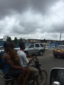 An Okada driver transporting 2 passengers on a cloudy day in Lagos